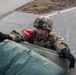 A soldier in MultiCam/OCP camouflage uniform is climbing over or peering over a barrier or vehicle edge, with green duffel bags or equipment visible in the foreground. The soldier is wearing a combat helmet with a mounted device, black tactical gloves, and shoulder patches including a red-bordered unit patch. The setting appears to be a paved military installation or training area.