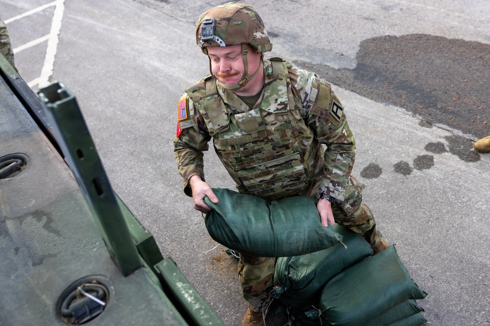 A U.S. Army soldier in full combat gear, including a camouflage uniform (OCP pattern), body armor, and helmet, is loading or stacking green sandbags onto or near a military vehicle. The soldier wears a U.S. flag patch on the right shoulder and what appears to be a captain's rank insignia. The activity suggests a flood control, defensive preparation, or disaster relief operation.