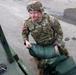 A U.S. Army soldier in full combat gear, including a camouflage uniform (OCP pattern), body armor, and helmet, is loading or stacking green sandbags onto or near a military vehicle. The soldier wears a U.S. flag patch on the right shoulder and what appears to be a captain's rank insignia. The activity suggests a flood control, defensive preparation, or disaster relief operation.