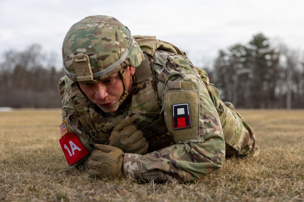 A U.S. Army soldier in full combat gear is performing a low crawl exercise on a grassy field. The soldier is wearing Operational Camouflage Pattern (OCP) uniform with a combat helmet, body armor, and tactical gloves. Visible unit patches and exercise markers are present on the uniform.