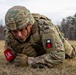A U.S. Army soldier in full combat gear is performing a low crawl exercise on a grassy field. The soldier is wearing Operational Camouflage Pattern (OCP) uniform with a combat helmet, body armor, and tactical gloves. Visible unit patches and exercise markers are present on the uniform.