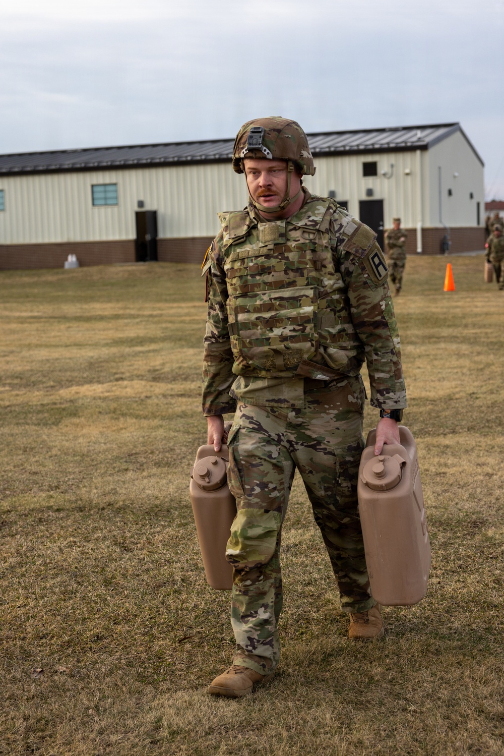 A U.S. Army soldier in full combat gear, including OCP (Operational Camouflage Pattern) uniform, body armor, and helmet, carries two tan military water/fuel canisters across a grassy field. The setting appears to be a military installation or training area, with a large building and orange traffic cone visible in the background. Other soldiers can be seen in the background, suggesting a unit-level training exercise or field activity.