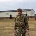A U.S. Army soldier in full combat gear, including OCP (Operational Camouflage Pattern) uniform, body armor, and helmet, carries two tan military water/fuel canisters across a grassy field. The setting appears to be a military installation or training area, with a large building and orange traffic cone visible in the background. Other soldiers can be seen in the background, suggesting a unit-level training exercise or field activity.