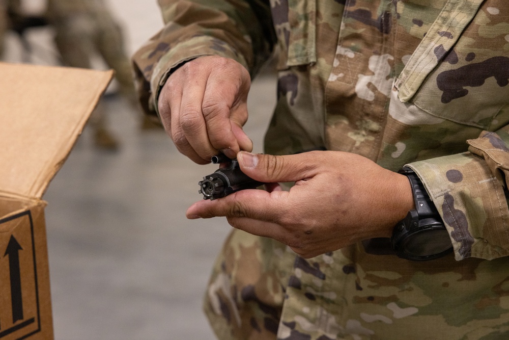 A person wearing military camouflage uniform (OCP/MultiCam pattern) is handling a small black mechanical component, likely a firearm muzzle device or flash hider. The individual is wearing a tactical watch on their left wrist and is positioned near a cardboard shipping box. The setting appears to be an indoor military supply or logistics environment.