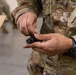 A person wearing military camouflage uniform (OCP/MultiCam pattern) is handling a small black mechanical component, likely a firearm muzzle device or flash hider. The individual is wearing a tactical watch on their left wrist and is positioned near a cardboard shipping box. The setting appears to be an indoor military supply or logistics environment.