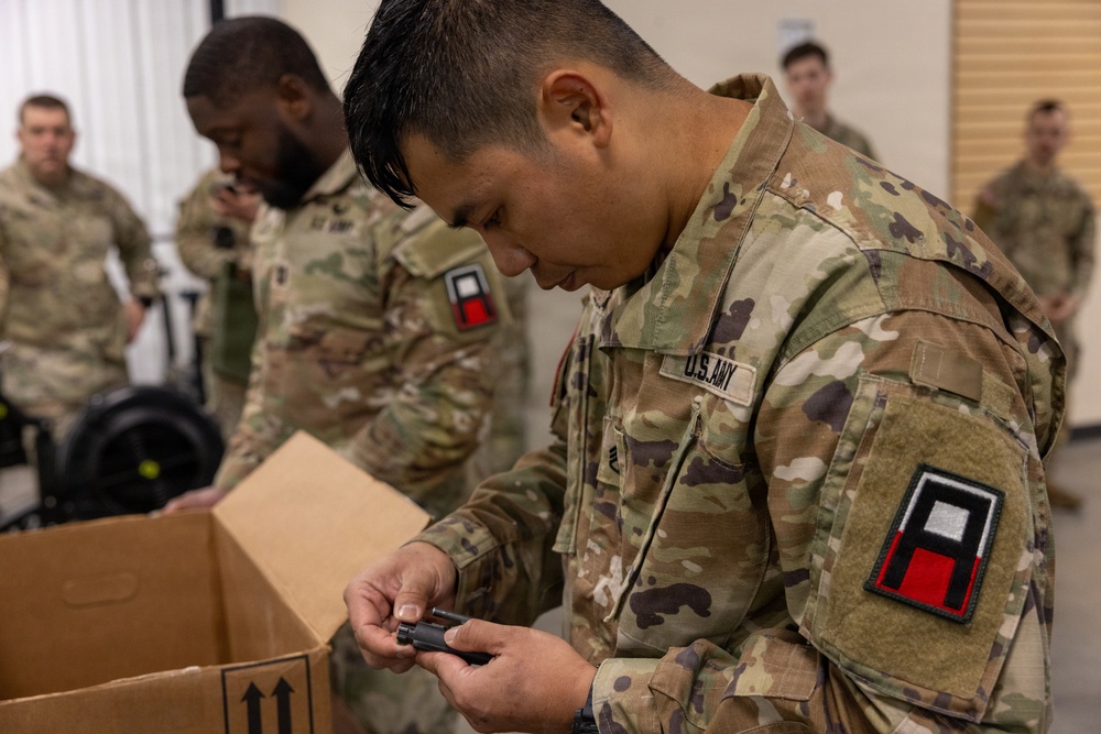 U.S. Army soldiers in Operational Camouflage Pattern (OCP) uniforms are engaged in what appears to be an equipment issue or inventory activity in an indoor facility. The foreground soldier is closely inspecting a small black component retrieved from a cardboard box, while other soldiers observe or assist in the background. The shoulder patch visible on the foreground soldier identifies affiliation with the First Army.