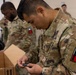 U.S. Army soldiers in Operational Camouflage Pattern (OCP) uniforms are engaged in what appears to be an equipment issue or inventory activity in an indoor facility. The foreground soldier is closely inspecting a small black component retrieved from a cardboard box, while other soldiers observe or assist in the background. The shoulder patch visible on the foreground soldier identifies affiliation with the First Army.