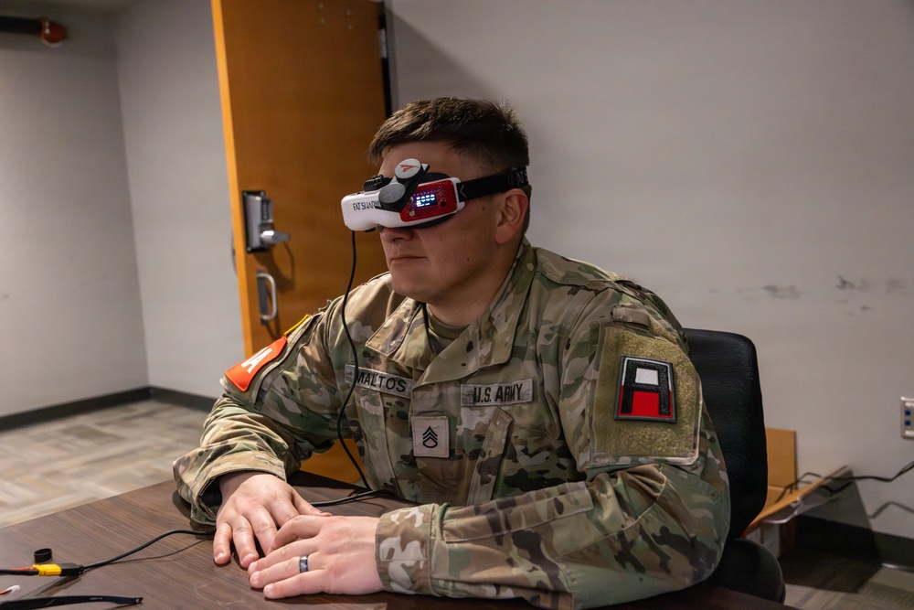A U.S. Army soldier wearing camouflage OCP (Operational Camouflage Pattern) uniform is seated at a desk using FPV (First Person View) goggles, identifiable as 'Fat Shark' brand. The soldier's name tape reads 'MALTOS' and rank insignia indicates the grade of Sergeant (E-5). The setting appears to be an indoor military facility or office environment, suggesting a drone or simulation training exercise.