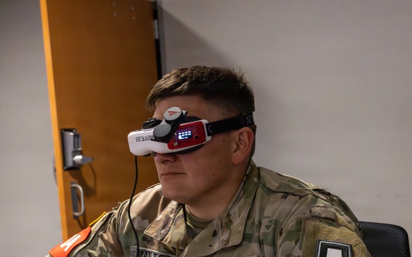 A U.S. Army soldier wearing camouflage OCP (Operational Camouflage Pattern) uniform is seated at a desk using FPV (First Person View) goggles, identifiable as 'Fat Shark' brand. The soldier's name tape reads 'MALTOS' and rank insignia indicates the grade of Sergeant (E-5). The setting appears to be an indoor military facility or office environment, suggesting a drone or simulation training exercise.