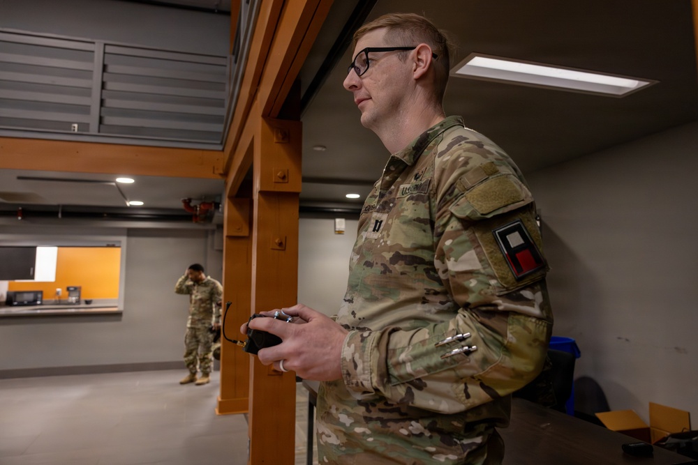 A U.S. Army soldier in OCP (Operational Camouflage Pattern) uniform is standing indoors holding what appears to be a remote control device, likely operating a drone or robotic system. A second soldier is visible in the background near what appears to be a service window or counter area. The setting appears to be an indoor facility or training environment.