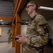 A U.S. Army soldier in OCP (Operational Camouflage Pattern) uniform is standing indoors holding what appears to be a remote control device, likely operating a drone or robotic system. A second soldier is visible in the background near what appears to be a service window or counter area. The setting appears to be an indoor facility or training environment.