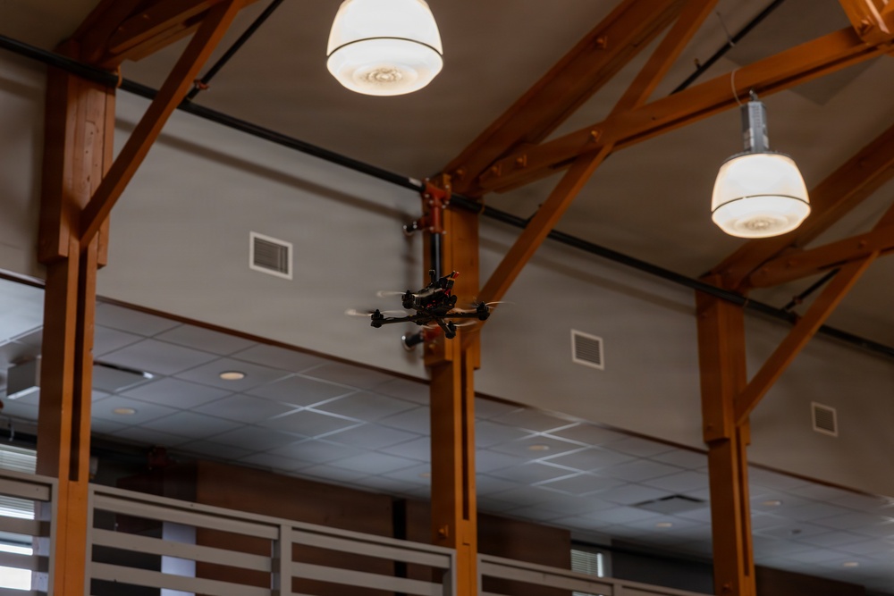 A small quadcopter drone is flying indoors within a large room featuring prominent wooden beam framework and pendant ceiling lights. The space appears to be a modern building interior, possibly a gymnasium, community center, or academic facility. The drone is captured mid-flight near the wooden structural beams.