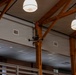 A small quadcopter drone is flying indoors within a large room featuring prominent wooden beam framework and pendant ceiling lights. The space appears to be a modern building interior, possibly a gymnasium, community center, or academic facility. The drone is captured mid-flight near the wooden structural beams.