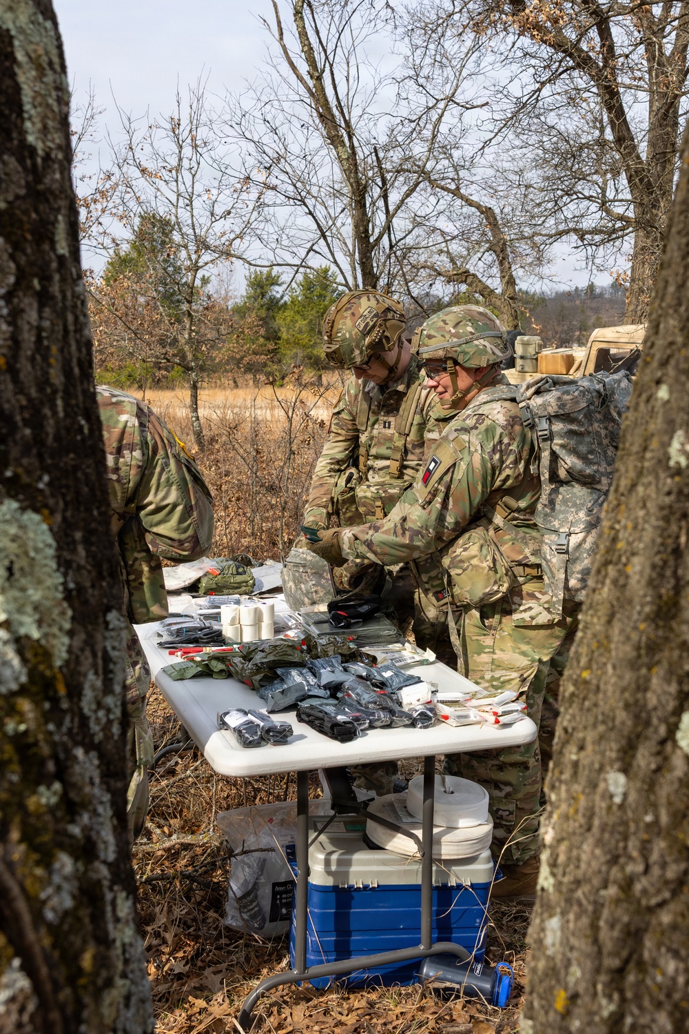 Several soldiers in MultiCam/OCP camouflage uniforms are gathered around a folding table in a wooded outdoor setting, examining and handling what appear to be medical or tactical supply packages. The table is covered with numerous sealed black pouches, white medical bandage rolls, and other packaged items, with a blue and white cooler stored underneath. The setting and equipment suggest a field training exercise or operational resupply activity.