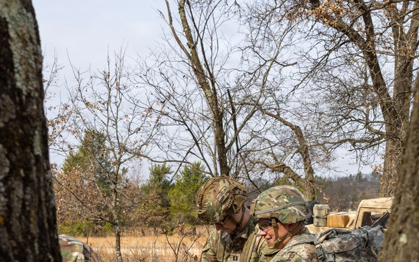 Several soldiers in MultiCam/OCP camouflage uniforms are gathered around a folding table in a wooded outdoor setting, examining and handling what appear to be medical or tactical supply packages. The table is covered with numerous sealed black pouches, white medical bandage rolls, and other packaged items, with a blue and white cooler stored underneath. The setting and equipment suggest a field training exercise or operational resupply activity.