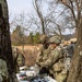 Several soldiers in MultiCam/OCP camouflage uniforms are gathered around a folding table in a wooded outdoor setting, examining and handling what appear to be medical or tactical supply packages. The table is covered with numerous sealed black pouches, white medical bandage rolls, and other packaged items, with a blue and white cooler stored underneath. The setting and equipment suggest a field training exercise or operational resupply activity.