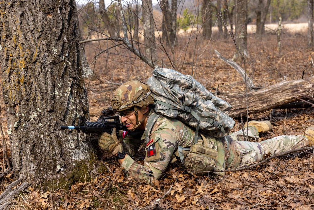 A soldier in U.S. Army MultiCam (OCP) camouflage uniform is in a prone firing position behind a large tree in a wooded area. The soldier is aiming a rifle equipped with a blue training barrel attachment, indicating this is a training exercise rather than combat. The soldier is carrying a large rucksack and wearing a tactical helmet.