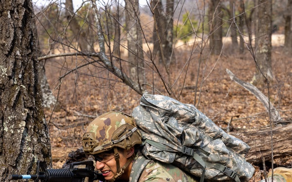 A soldier in U.S. Army MultiCam (OCP) camouflage uniform is in a prone firing position behind a large tree in a wooded area. The soldier is aiming a rifle equipped with a blue training barrel attachment, indicating this is a training exercise rather than combat. The soldier is carrying a large rucksack and wearing a tactical helmet.