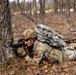 A soldier in U.S. Army MultiCam (OCP) camouflage uniform is in a prone firing position behind a large tree in a wooded area. The soldier is aiming a rifle equipped with a blue training barrel attachment, indicating this is a training exercise rather than combat. The soldier is carrying a large rucksack and wearing a tactical helmet.