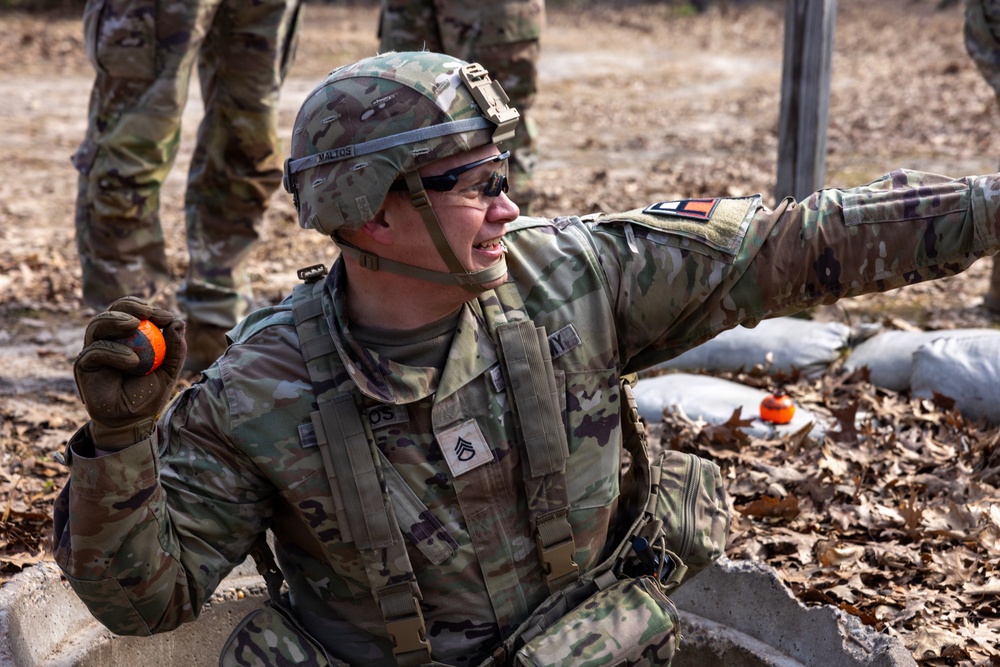 A U.S. Army Staff Sergeant in full combat gear is crouched in what appears to be a training exercise, holding an orange practice grenade in one hand while pointing with the other. The soldier is wearing Operational Camouflage Pattern (OCP) uniform, a combat helmet with the name 'MALTOS' visible on the band, and tactical sunglasses. The setting appears to be an outdoor training range with sandbags and leaf-covered ground visible.