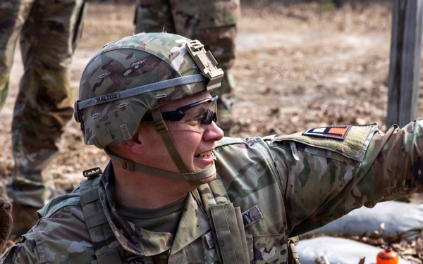 A U.S. Army Staff Sergeant in full combat gear is crouched in what appears to be a training exercise, holding an orange practice grenade in one hand while pointing with the other. The soldier is wearing Operational Camouflage Pattern (OCP) uniform, a combat helmet with the name 'MALTOS' visible on the band, and tactical sunglasses. The setting appears to be an outdoor training range with sandbags and leaf-covered ground visible.