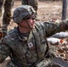 A U.S. Army Staff Sergeant in full combat gear is crouched in what appears to be a training exercise, holding an orange practice grenade in one hand while pointing with the other. The soldier is wearing Operational Camouflage Pattern (OCP) uniform, a combat helmet with the name 'MALTOS' visible on the band, and tactical sunglasses. The setting appears to be an outdoor training range with sandbags and leaf-covered ground visible.