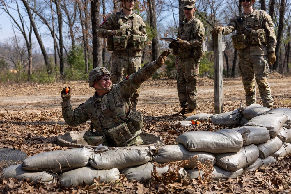 A U.S. Army soldier in a foxhole or fighting position surrounded by sandbags appears to be practicing grenade throwing during a training exercise. Three other soldiers in camouflage uniforms stand nearby observing and taking notes, suggesting they are evaluators or instructors. The setting is an outdoor training area with bare trees and dry leaf-covered ground.