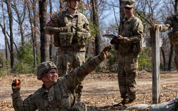 A U.S. Army soldier in a foxhole or fighting position surrounded by sandbags appears to be practicing grenade throwing during a training exercise. Three other soldiers in camouflage uniforms stand nearby observing and taking notes, suggesting they are evaluators or instructors. The setting is an outdoor training area with bare trees and dry leaf-covered ground.