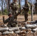 A U.S. Army soldier in a foxhole or fighting position surrounded by sandbags appears to be practicing grenade throwing during a training exercise. Three other soldiers in camouflage uniforms stand nearby observing and taking notes, suggesting they are evaluators or instructors. The setting is an outdoor training area with bare trees and dry leaf-covered ground.