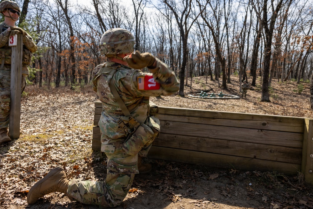 A U.S. Army soldier in full OCP (Operational Camouflage Pattern) uniform is kneeling behind a wooden barrier and appears to be throwing a practice grenade during a training exercise. A second soldier wearing sunglasses stands near a post marked with the number '3', likely serving as an observer or evaluator. The setting is an outdoor wooded training range with leaf-covered ground.