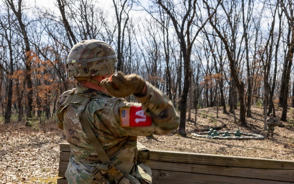A U.S. Army soldier in full OCP (Operational Camouflage Pattern) uniform is kneeling behind a wooden barrier and appears to be throwing a practice grenade during a training exercise. A second soldier wearing sunglasses stands near a post marked with the number '3', likely serving as an observer or evaluator. The setting is an outdoor wooded training range with leaf-covered ground.