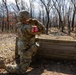A U.S. Army soldier in full OCP (Operational Camouflage Pattern) uniform is kneeling behind a wooden barrier and appears to be throwing a practice grenade during a training exercise. A second soldier wearing sunglasses stands near a post marked with the number '3', likely serving as an observer or evaluator. The setting is an outdoor wooded training range with leaf-covered ground.