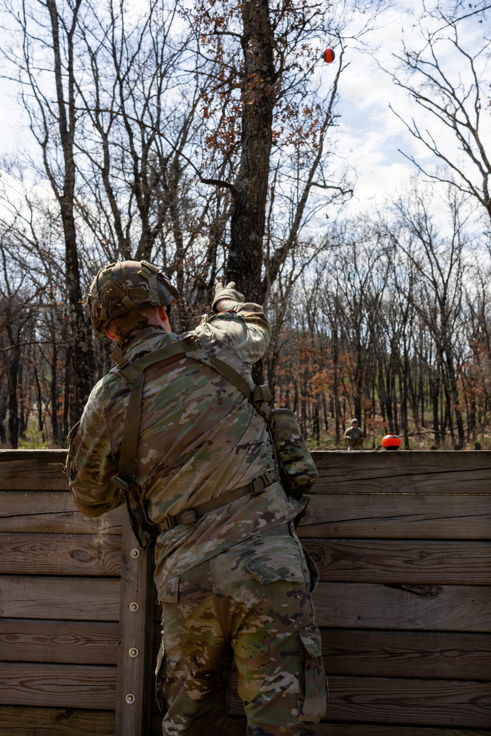 A soldier in full MultiCam/OCP camouflage uniform and tactical gear is throwing what appears to be a practice grenade (orange/red training grenade) over a wooden barrier wall in a wooded training environment. The grenade is visibly airborne in the upper portion of the frame. A second soldier is faintly visible in the background beyond the wall.