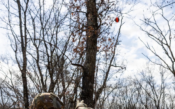 A soldier in full MultiCam/OCP camouflage uniform and tactical gear is throwing what appears to be a practice grenade (orange/red training grenade) over a wooden barrier wall in a wooded training environment. The grenade is visibly airborne in the upper portion of the frame. A second soldier is faintly visible in the background beyond the wall.