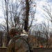 A soldier in full MultiCam/OCP camouflage uniform and tactical gear is throwing what appears to be a practice grenade (orange/red training grenade) over a wooden barrier wall in a wooded training environment. The grenade is visibly airborne in the upper portion of the frame. A second soldier is faintly visible in the background beyond the wall.