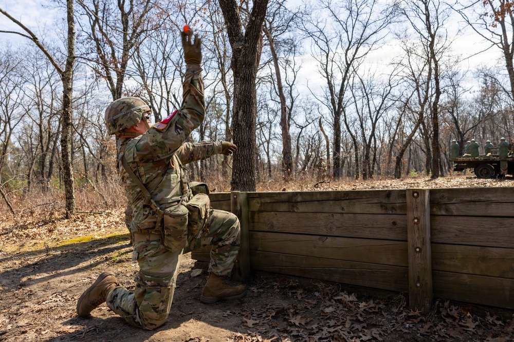 A U.S. Army soldier in Operational Camouflage Pattern (OCP) uniform is kneeling behind a wooden barrier and throwing what appears to be a practice/training grenade (orange colored). The setting is a wooded training area with leafless trees, suggesting late autumn or winter conditions. Additional military personnel and equipment are visible in the background.