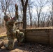 A U.S. Army soldier in Operational Camouflage Pattern (OCP) uniform is kneeling behind a wooden barrier and throwing what appears to be a practice/training grenade (orange colored). The setting is a wooded training area with leafless trees, suggesting late autumn or winter conditions. Additional military personnel and equipment are visible in the background.