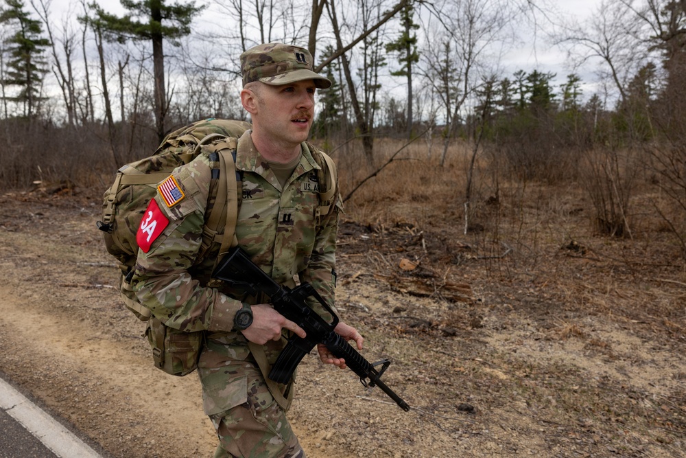 A U.S. Army soldier in Operational Camouflage Pattern (OCP) uniform is conducting a ruck march along a road in a wooded area during what appears to be late autumn or early spring. The soldier is carrying a large rucksack and holding an M4/M16-style rifle while moving at a brisk pace. Visible insignia indicate U.S. Army affiliation, an officer rank (Captain/First Lieutenant), and a red unit patch.