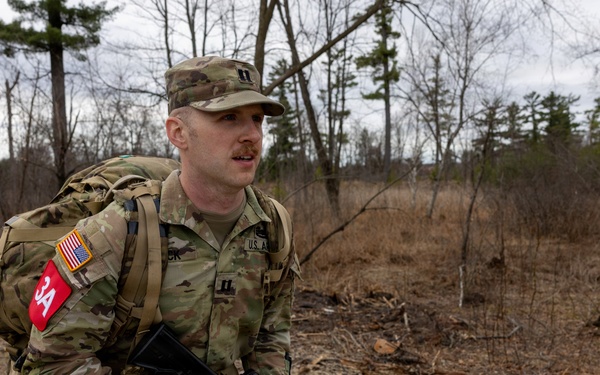 A U.S. Army soldier in Operational Camouflage Pattern (OCP) uniform is conducting a ruck march along a road in a wooded area during what appears to be late autumn or early spring. The soldier is carrying a large rucksack and holding an M4/M16-style rifle while moving at a brisk pace. Visible insignia indicate U.S. Army affiliation, an officer rank (Captain/First Lieutenant), and a red unit patch.