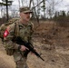 A U.S. Army soldier in Operational Camouflage Pattern (OCP) uniform is conducting a ruck march along a road in a wooded area during what appears to be late autumn or early spring. The soldier is carrying a large rucksack and holding an M4/M16-style rifle while moving at a brisk pace. Visible insignia indicate U.S. Army affiliation, an officer rank (Captain/First Lieutenant), and a red unit patch.