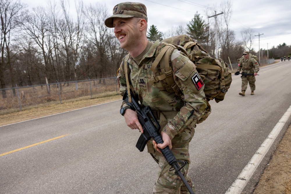 A smiling U.S. Army soldier in OCP (Operational Camouflage Pattern) uniform is conducting a ruck march along a paved road, carrying a large rucksack and an M4/M16-style rifle. At least one additional soldier is visible in the background performing the same activity. The setting appears to be a military installation or training area during late fall or early winter based on the bare deciduous trees.