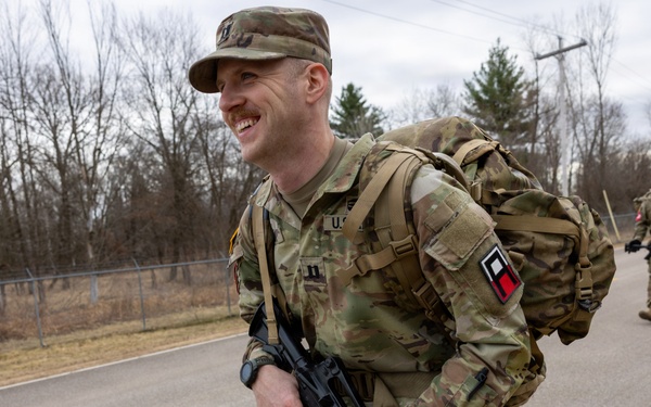 A smiling U.S. Army soldier in OCP (Operational Camouflage Pattern) uniform is conducting a ruck march along a paved road, carrying a large rucksack and an M4/M16-style rifle. At least one additional soldier is visible in the background performing the same activity. The setting appears to be a military installation or training area during late fall or early winter based on the bare deciduous trees.