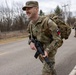 A smiling U.S. Army soldier in OCP (Operational Camouflage Pattern) uniform is conducting a ruck march along a paved road, carrying a large rucksack and an M4/M16-style rifle. At least one additional soldier is visible in the background performing the same activity. The setting appears to be a military installation or training area during late fall or early winter based on the bare deciduous trees.
