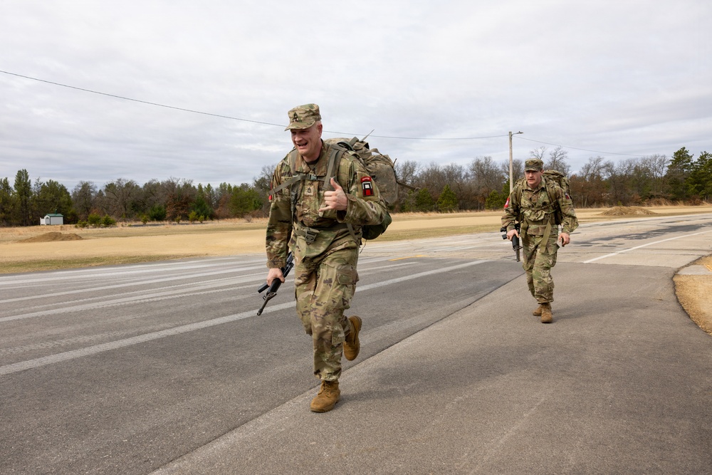 Two U.S. Army soldiers in OCP (Operational Camouflage Pattern) uniforms are conducting a ruck march along a paved road in what appears to be a military installation or training area. The lead soldier is running and giving a thumbs-up gesture while carrying a rifle and a loaded rucksack. The setting appears to be a cold, overcast day in a rural or semi-rural environment.
