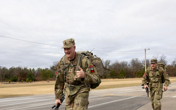 Two U.S. Army soldiers in OCP (Operational Camouflage Pattern) uniforms are conducting a ruck march along a paved road in what appears to be a military installation or training area. The lead soldier is running and giving a thumbs-up gesture while carrying a rifle and a loaded rucksack. The setting appears to be a cold, overcast day in a rural or semi-rural environment.