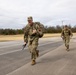 Two U.S. Army soldiers in OCP (Operational Camouflage Pattern) uniforms are conducting a ruck march along a paved road in what appears to be a military installation or training area. The lead soldier is running and giving a thumbs-up gesture while carrying a rifle and a loaded rucksack. The setting appears to be a cold, overcast day in a rural or semi-rural environment.