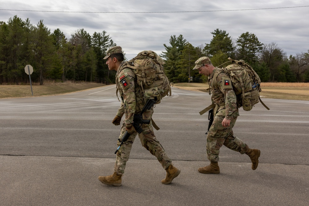 Two military personnel in full combat gear are conducting a road march along a paved road in what appears to be a training installation. Both soldiers are wearing MultiCam (OCP) camouflage uniforms, carrying large rucksacks, and are armed with rifles. The setting appears to be a military base or training area during late fall or early winter based on the vegetation and overcast sky.