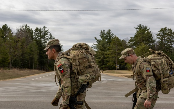 Two military personnel in full combat gear are conducting a road march along a paved road in what appears to be a training installation. Both soldiers are wearing MultiCam (OCP) camouflage uniforms, carrying large rucksacks, and are armed with rifles. The setting appears to be a military base or training area during late fall or early winter based on the vegetation and overcast sky.