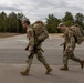 Two military personnel in full combat gear are conducting a road march along a paved road in what appears to be a training installation. Both soldiers are wearing MultiCam (OCP) camouflage uniforms, carrying large rucksacks, and are armed with rifles. The setting appears to be a military base or training area during late fall or early winter based on the vegetation and overcast sky.