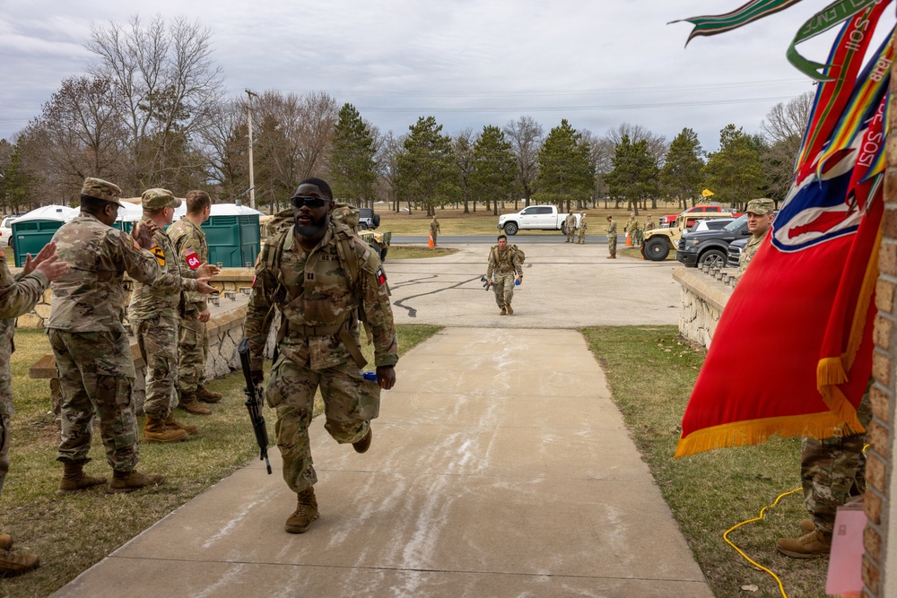Military personnel in Army Combat Uniforms (ACU/OCP pattern) are participating in what appears to be a ruck march or physical fitness event at a military installation. A lead soldier carrying a rucksack and rifle is walking along a paved pathway while fellow soldiers line the sides, appearing to offer encouragement. A red and gold military unit guidon flag is visible on the right side of the frame.